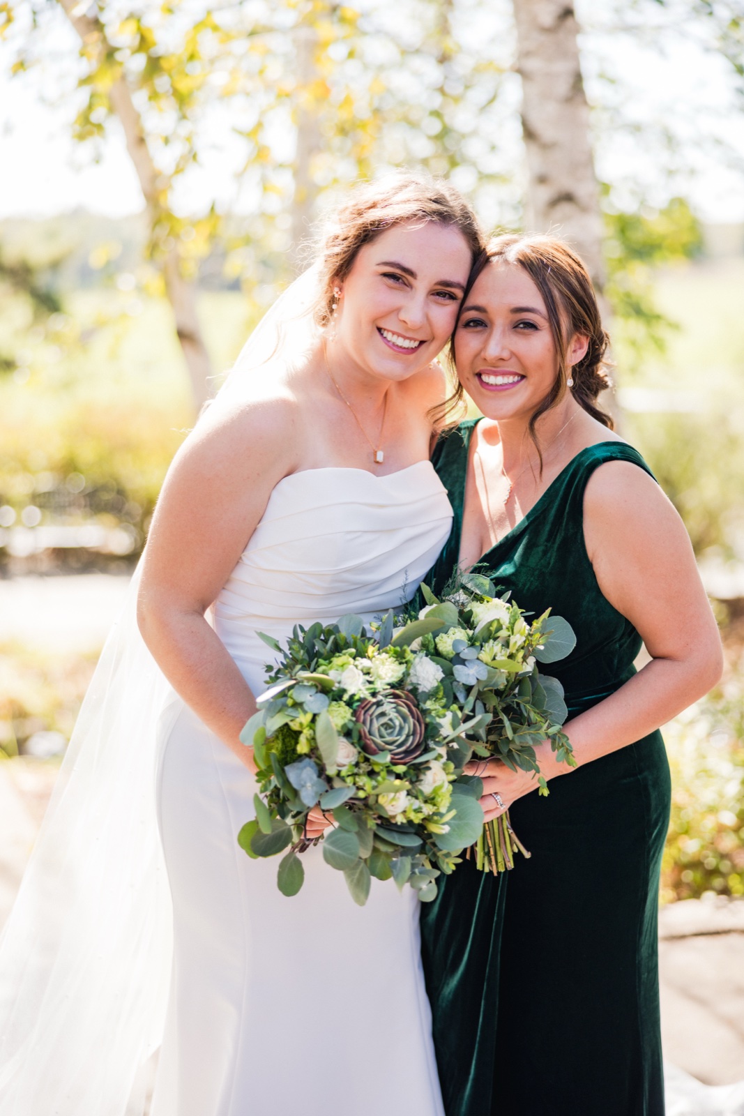 The bride and a bridesmaid in an emerald velvet dress lean cheek-to-cheek and smile at the camera, the bride holding a lush greenery-and-succulent bouquet under backlit trees — Tim Larsen Photography, Brainerd Lakes MN