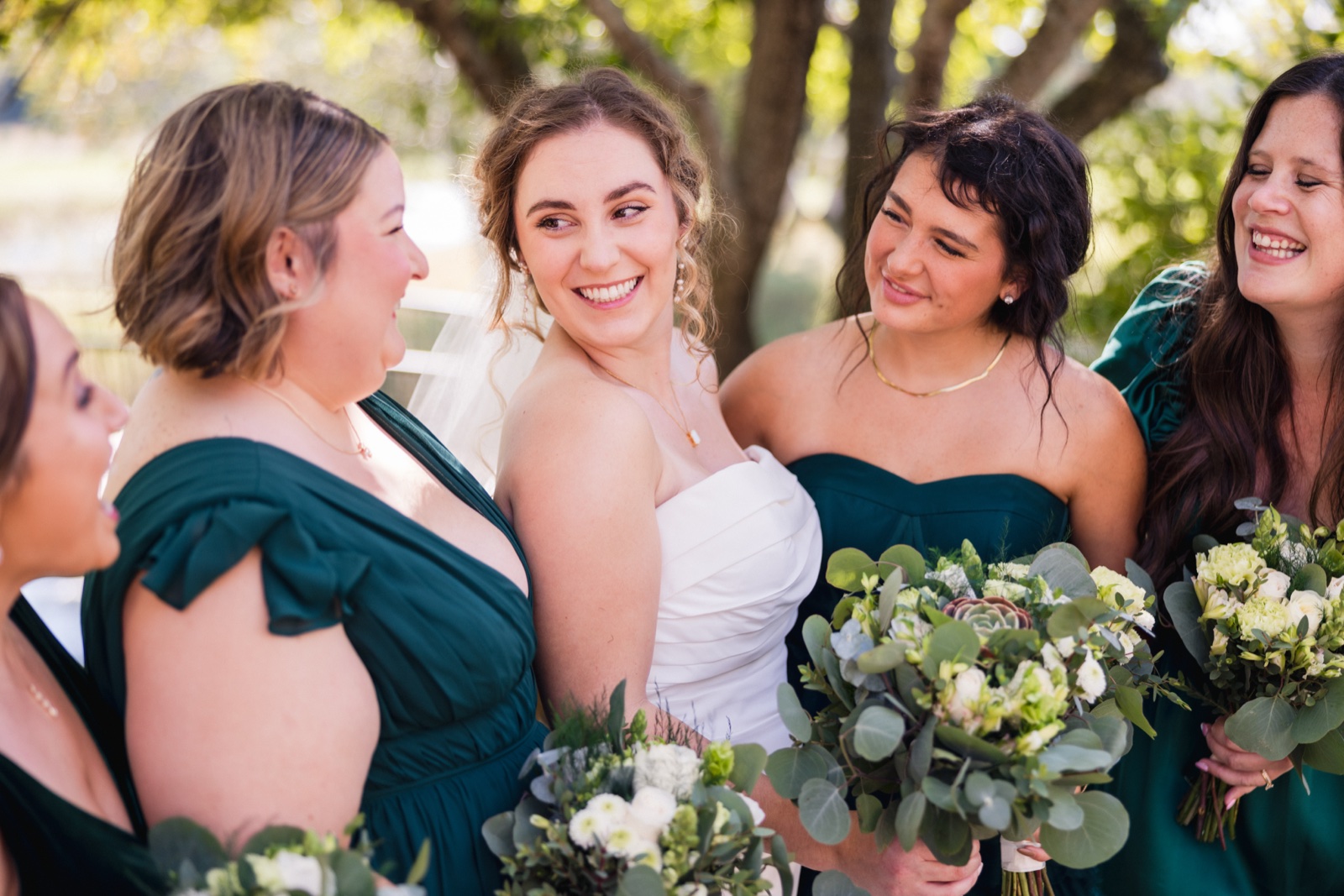 The bride laughs at the center of her bridesmaids in emerald dresses, all holding eucalyptus and white floral bouquets under dappled tree light — Tim Larsen Photography, Brainerd Lakes MN