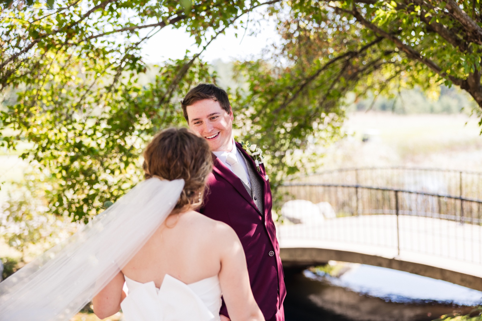 The groom in a burgundy suit smiles at his bride, seen from behind with her veil draped over her shoulder, on a garden bridge framed by overhanging leaves — Tim Larsen Photography, Brainerd Lakes MN