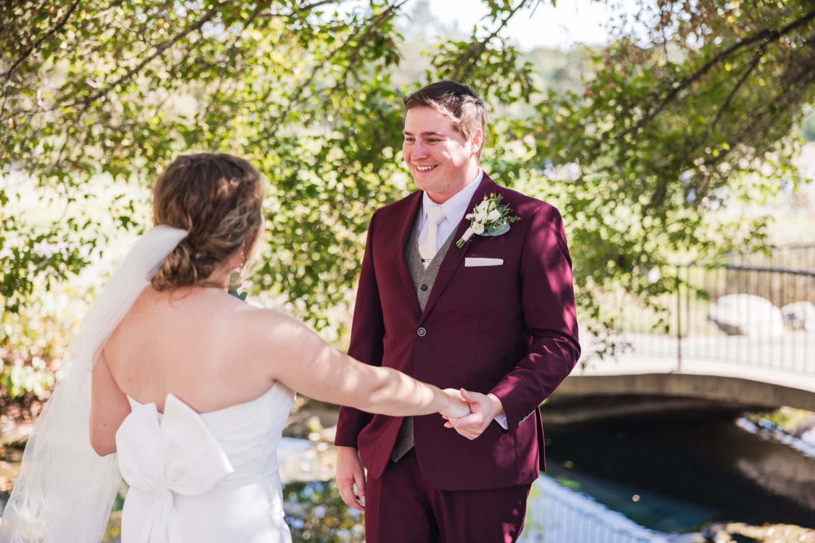 The groom in a burgundy suit smiles broadly as he holds the bride's hand during their first look on a garden bridge — Tim Larsen Photography, Brainerd Lakes MN