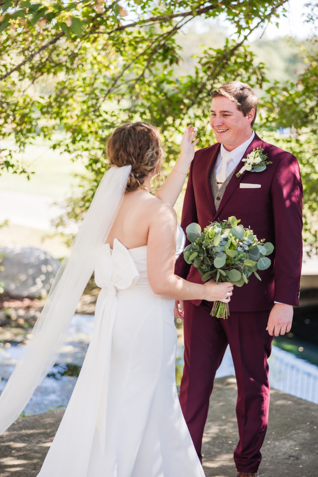 The bride reaches toward the groom's face as he smiles at her during their first look beneath a leafy tree, her long veil and his burgundy suit holding her bouquet visible in the frame — Tim Larsen Photography, Brainerd Lakes MN