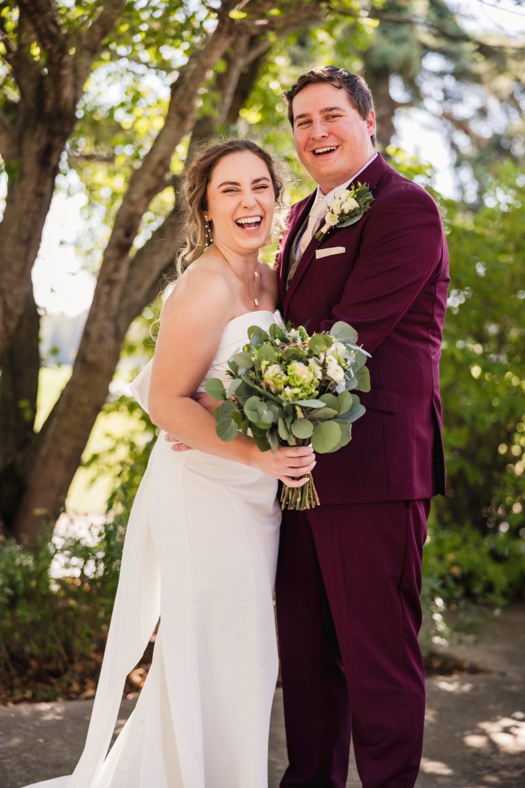 The bride and groom laugh openly while standing close together on a tree-shaded path, the bride holding a green-and-white bouquet against her ivory gown and the groom in a burgundy suit — Tim Larsen Photography, Brainerd Lakes MN