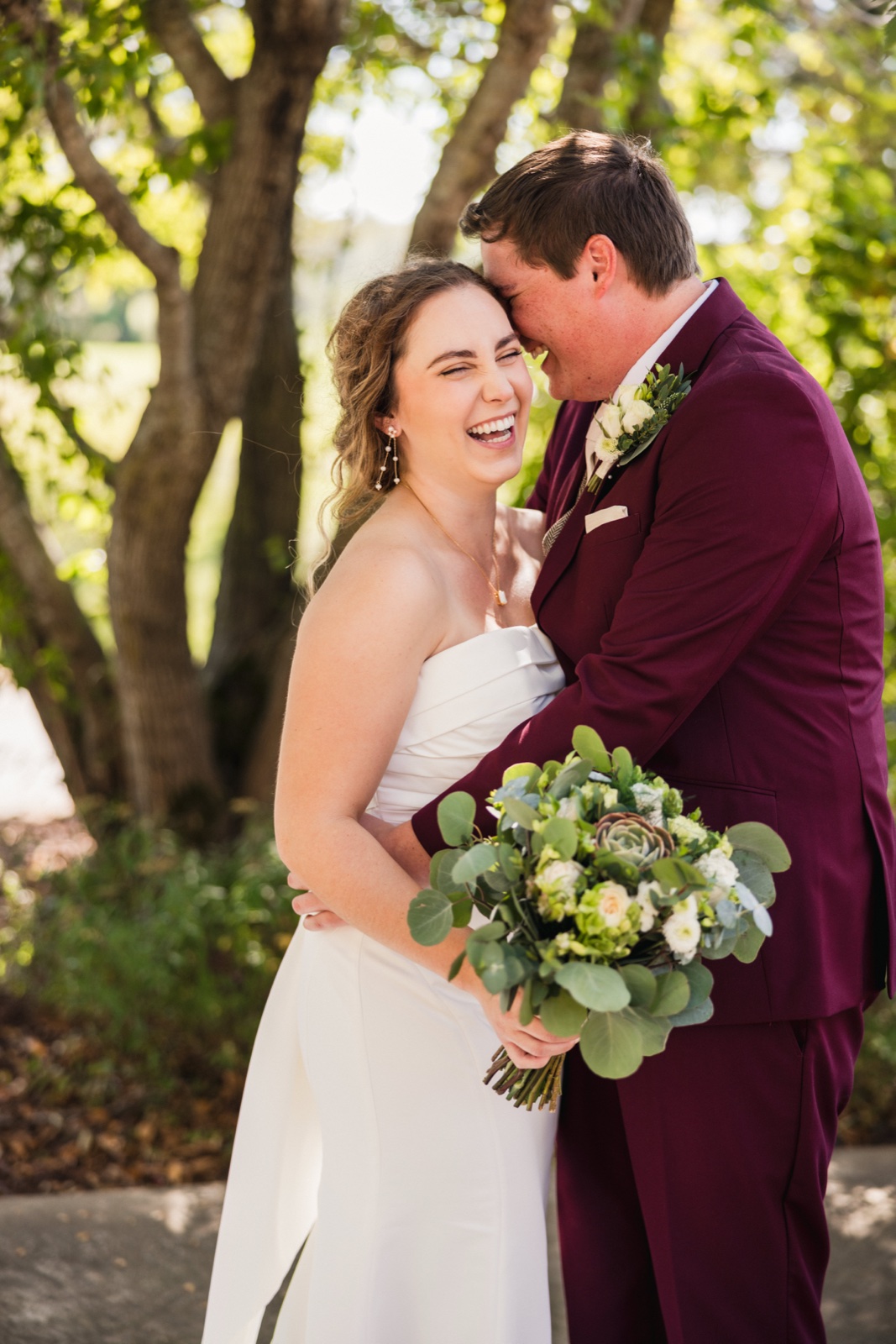 The bride laughs openly as the groom in a burgundy suit leans his forehead into hers, her bouquet of greenery and pale florals held between them under a tree canopy — Tim Larsen Photography, Brainerd Lakes MN