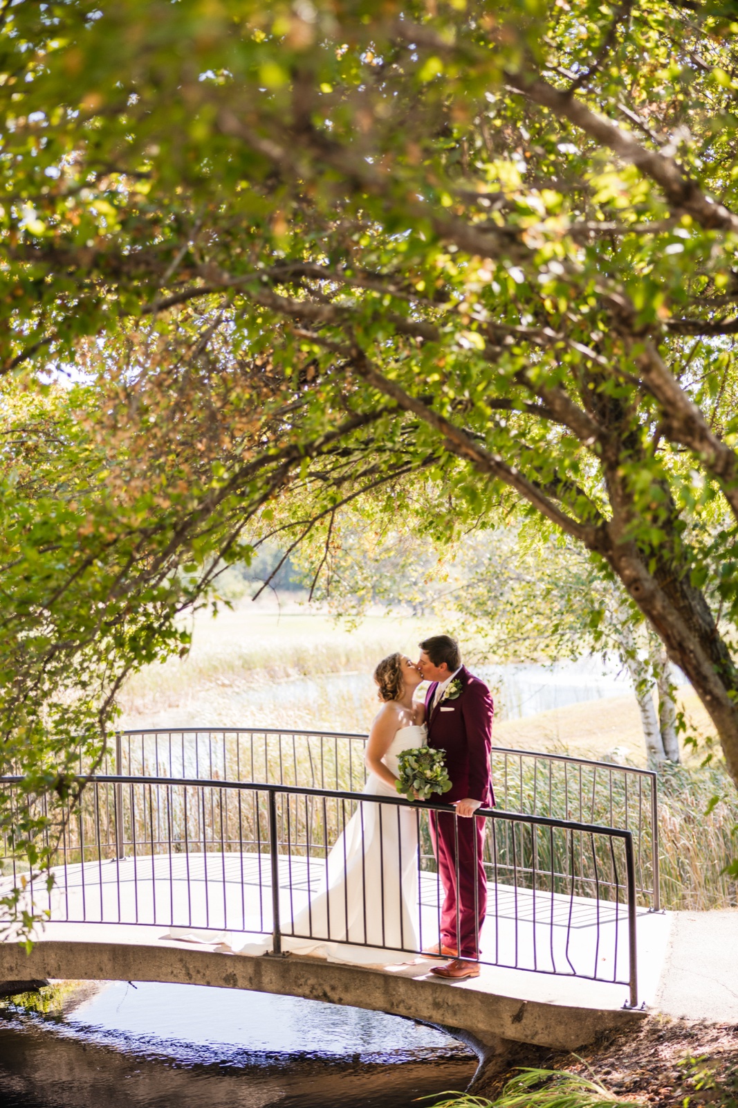The bride and groom share a kiss on a small white arched footbridge surrounded by tall trees and lakeside grasses on the Breezy Point Resort grounds — Tim Larsen Photography, Brainerd Lakes MN