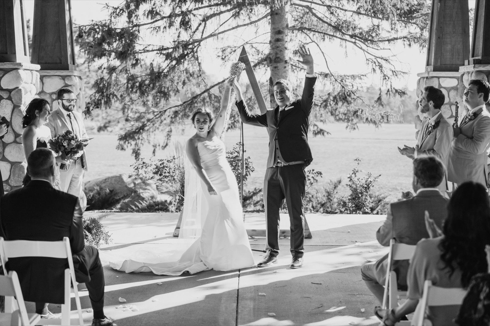 The bride and groom raise their arms in celebration just after their ceremony, framed by stone pillars and pine branches as guests applaud from their seats — Tim Larsen Photography, Brainerd Lakes MN