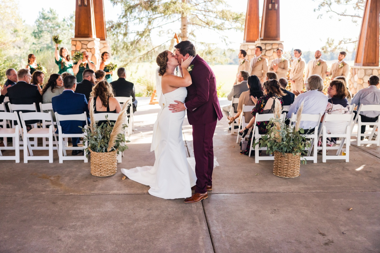 The bride and groom kiss in the center aisle of an outdoor stone-pillared ceremony pavilion as their bridal party and seated guests look on — Tim Larsen Photography, Brainerd Lakes MN