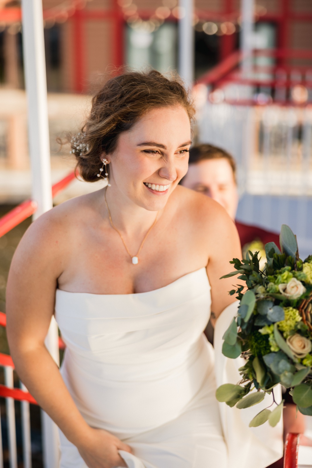 The bride laughs broadly while stepping onto the Breezy Belle paddleboat in golden evening light, holding a lush green-and-white bouquet — Tim Larsen Photography, Brainerd Lakes MN