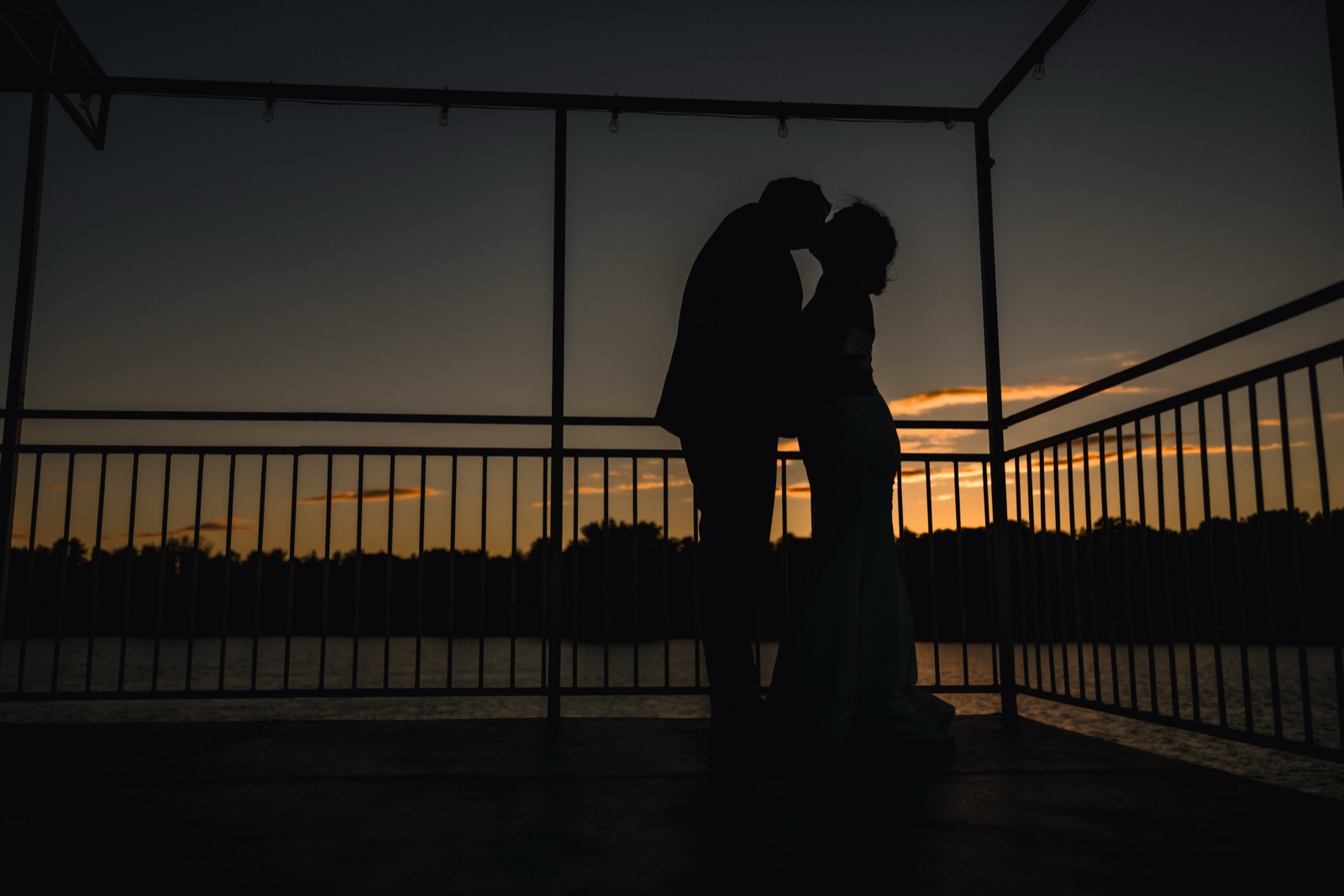 The bride and groom kiss in silhouette beneath a string-lit dock pavilion as the sky burns orange over the lake at dusk — Tim Larsen Photography, Brainerd Lakes MN