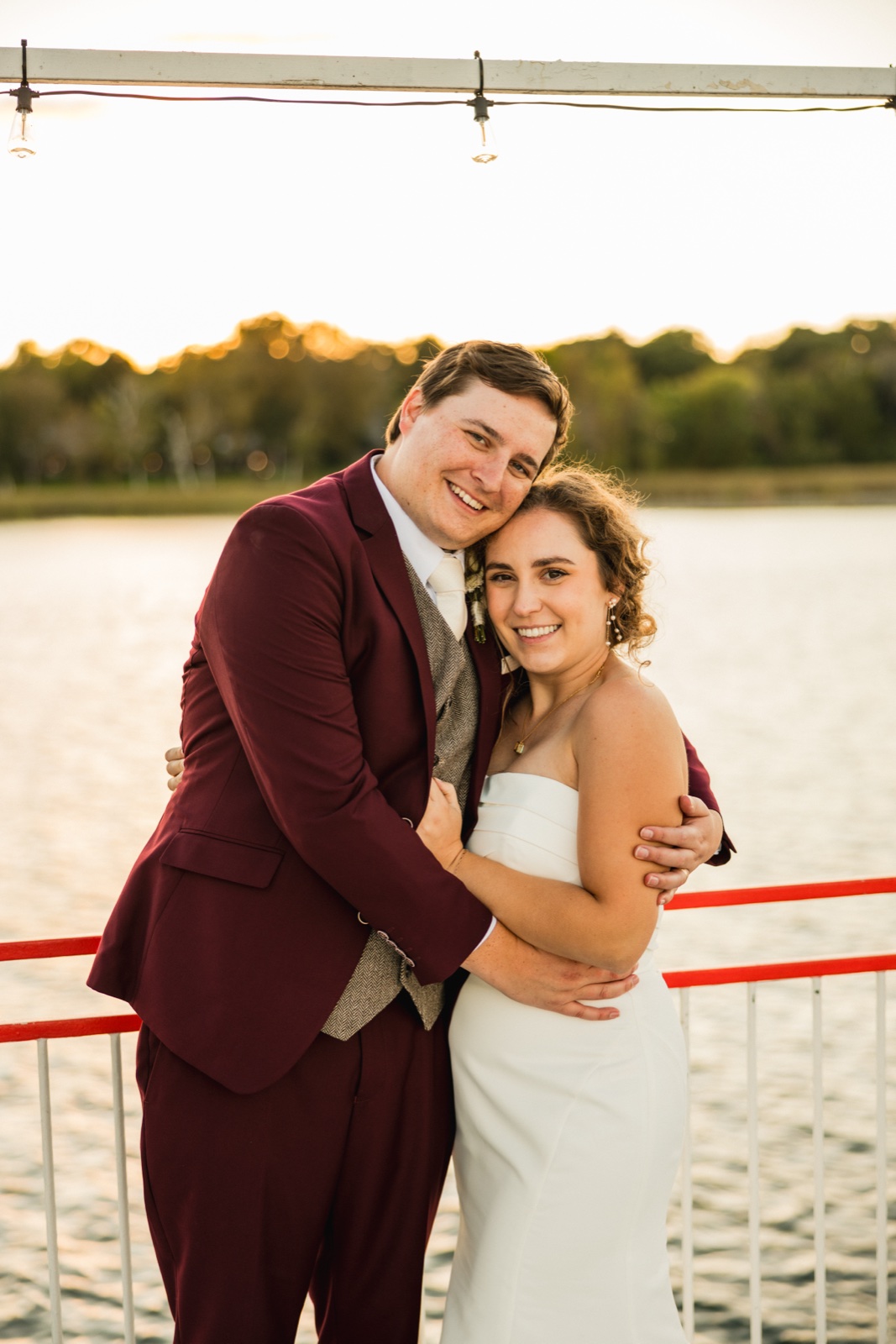 The bride and groom lean their heads together and smile at the camera while embracing on the deck of a boat at sunset, with a lake and string lights visible behind them — Tim Larsen Photography, Brainerd Lakes MN