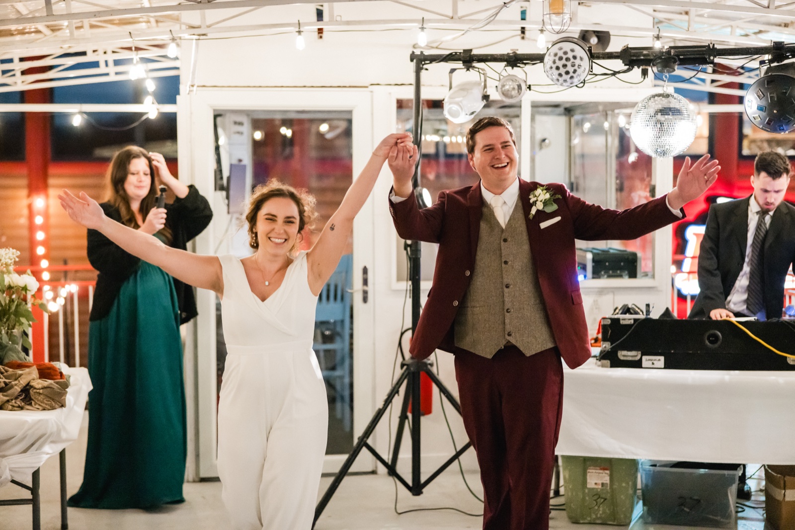 The bride in a white jumpsuit and the groom in a burgundy suit raise their arms in celebration as they enter the reception under string lights and a disco ball — Tim Larsen Photography, Brainerd Lakes MN