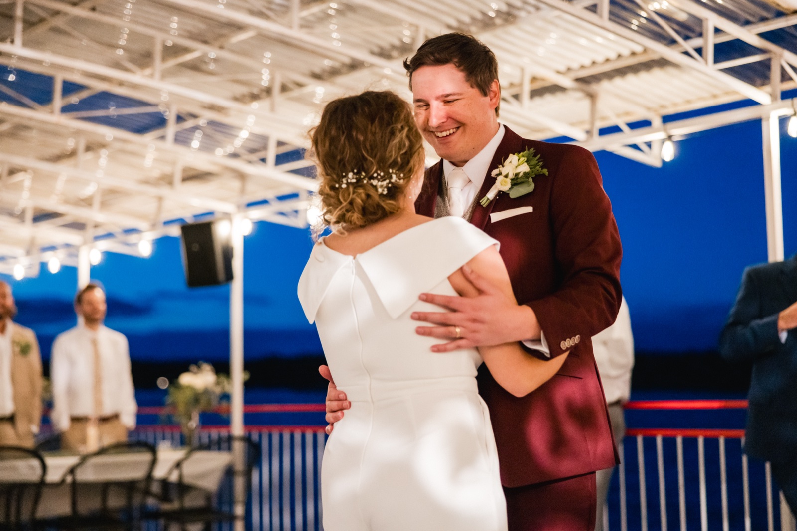 The groom in a burgundy suit grins broadly over the bride's shoulder during their first dance under a string-lit pavilion at dusk — Tim Larsen Photography, Brainerd Lakes MN
