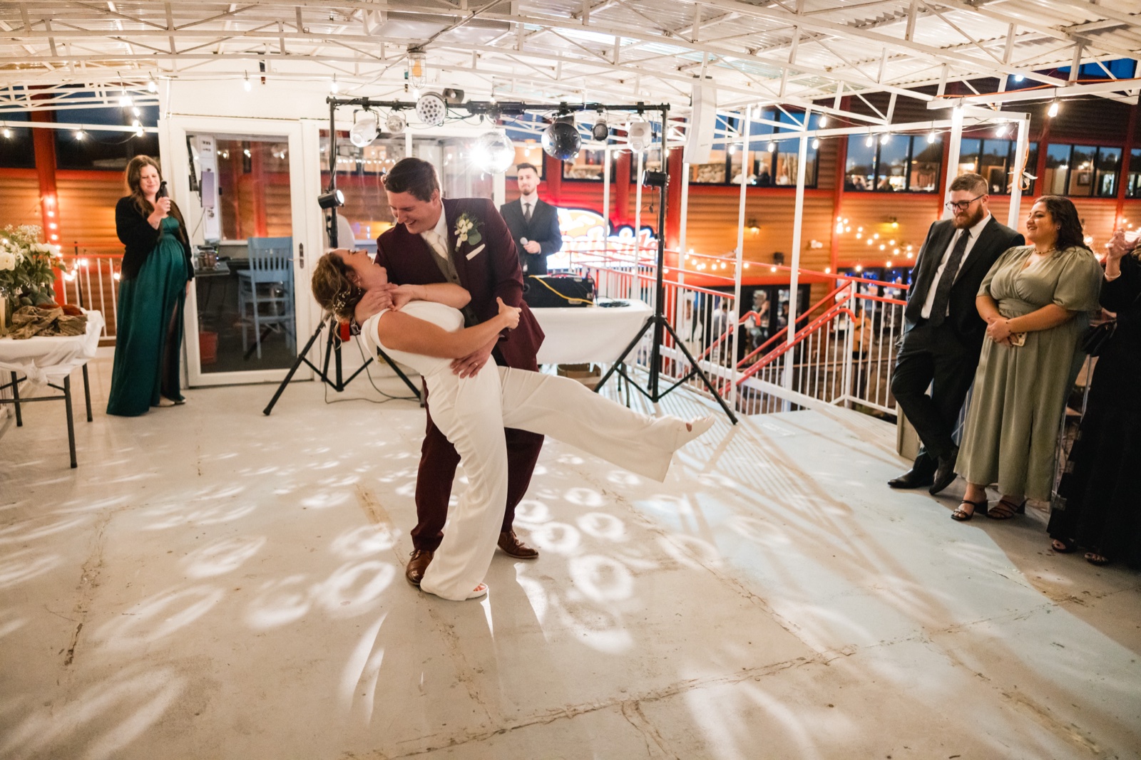 The groom dips the bride on a dance floor patterned with circular gobo lights as the bridal party and guests watch from the railing of an open-air pavilion at night — Tim Larsen Photography, Brainerd Lakes MN