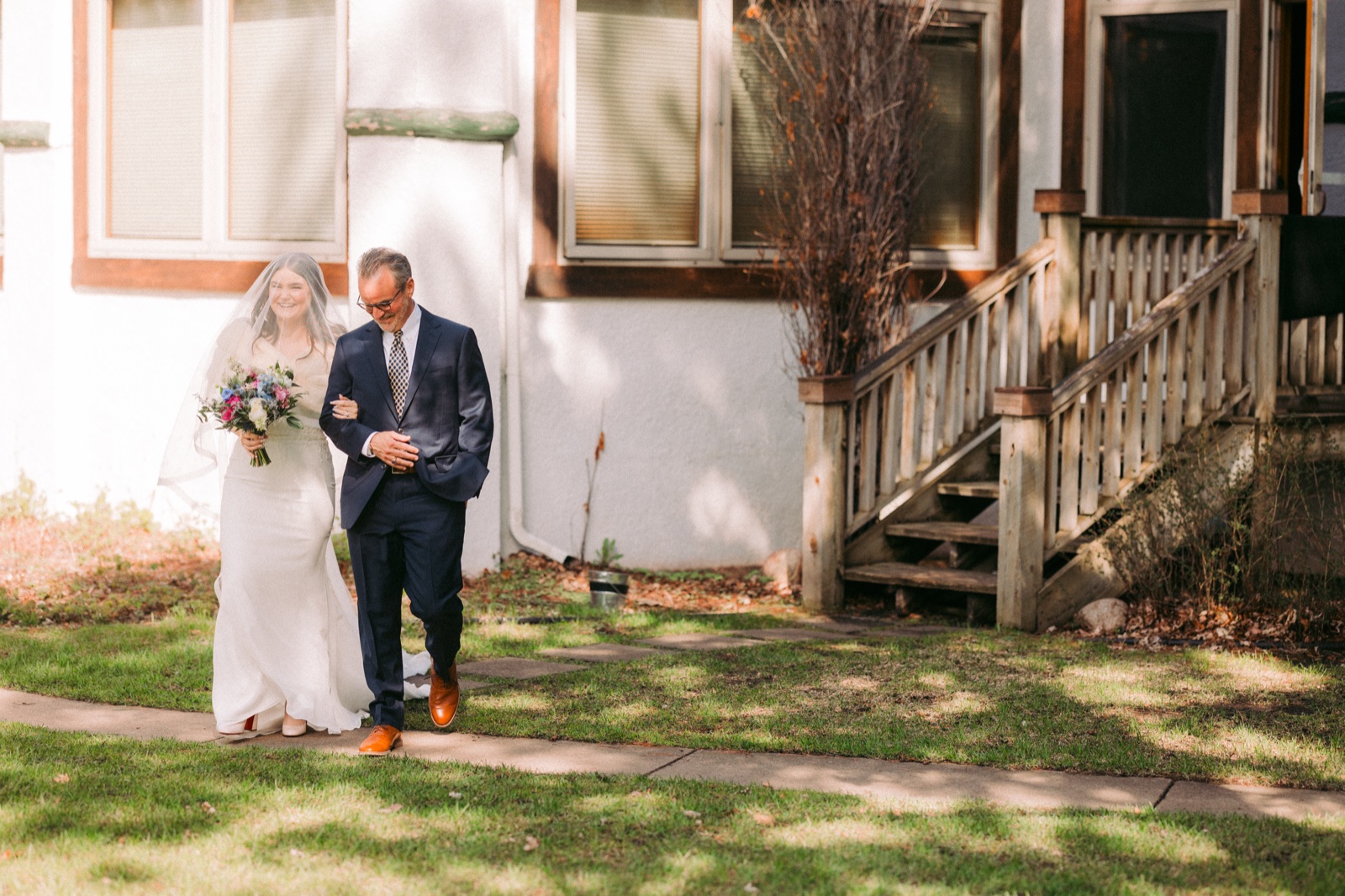 The bride, smiling broadly behind her veil and holding a colorful bouquet, walks down a stone path with her father along the side of the lodge — Tim Larsen Photography, Brainerd Lakes MN