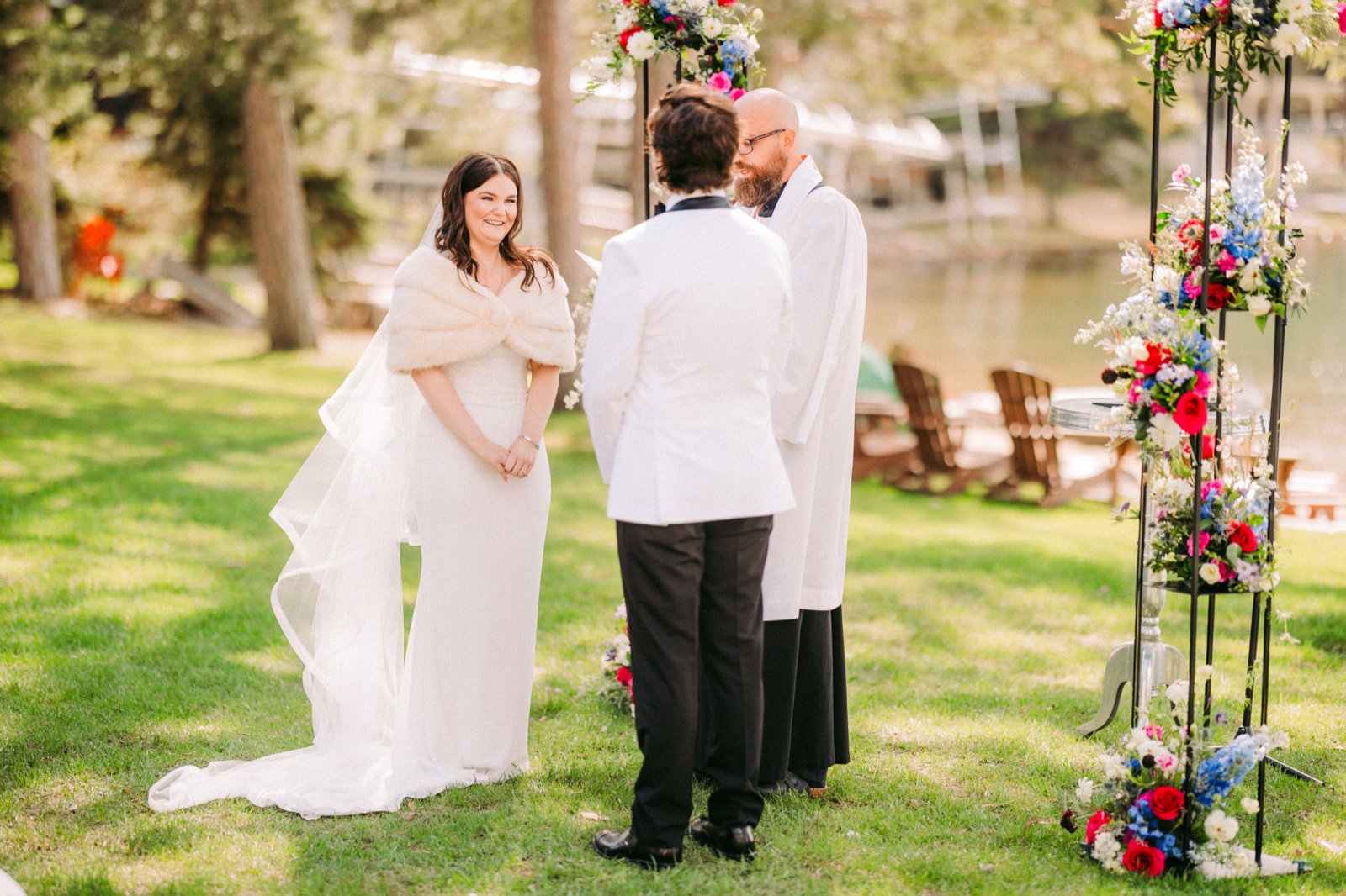 The bride in a fur stole and long veil smiles toward the groom during the lakeside ceremony, with the officiant beside him and a vivid floral arch framing the scene — Tim Larsen Photography, Brainerd Lakes MN