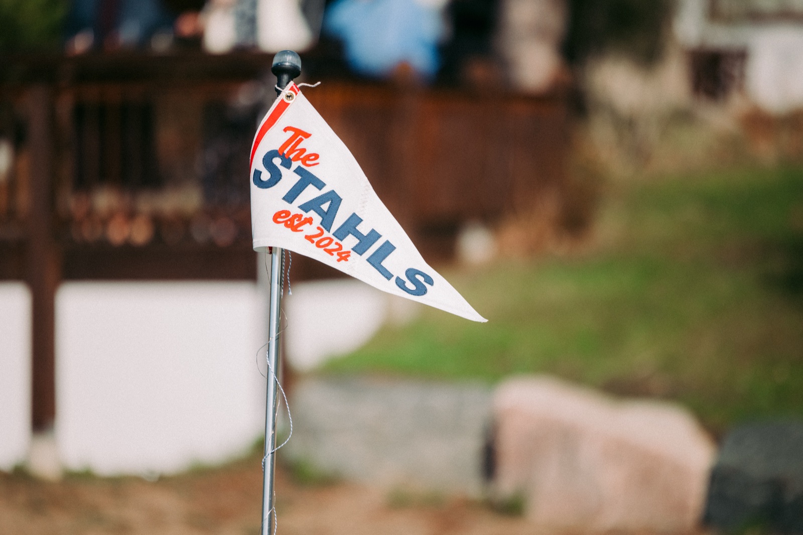 A small white pennant flag reading 'The Stahls est. 2024' in red and navy lettering stands in front of a softly blurred lodge railing — Tim Larsen Photography, Brainerd Lakes MN