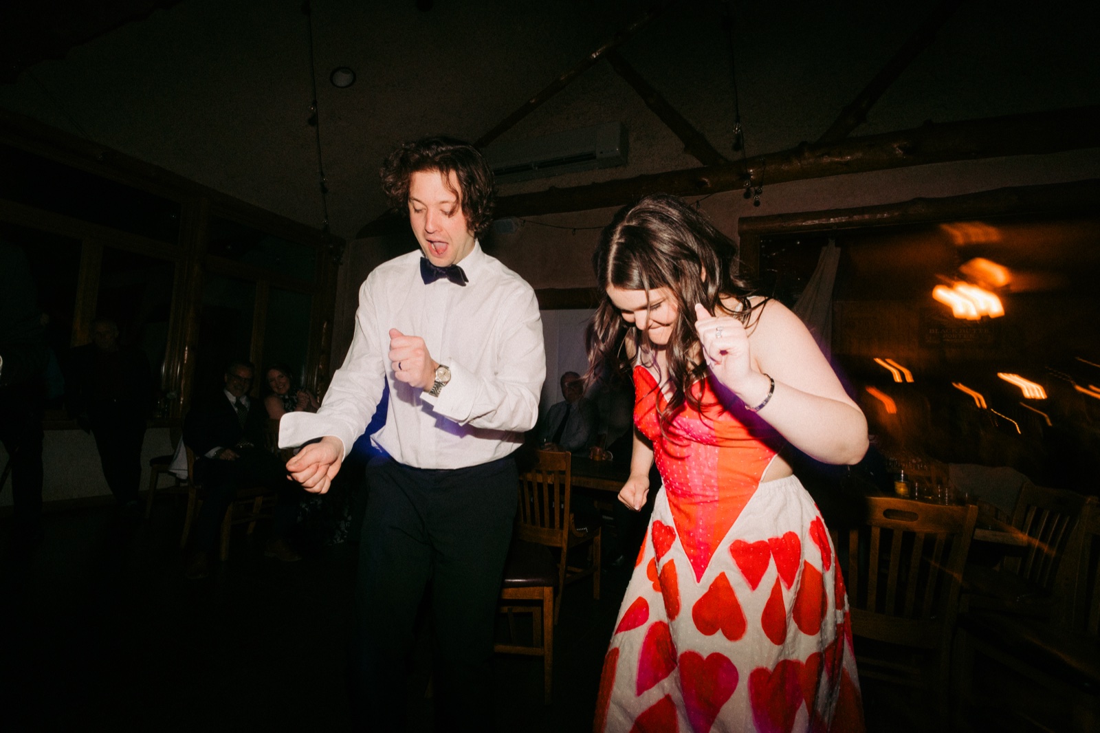 The bride in a heart-printed dress and the groom in a bow tie dance with loose, playful energy under the wood-beamed ceiling of the reception hall — Tim Larsen Photography, Brainerd Lakes MN