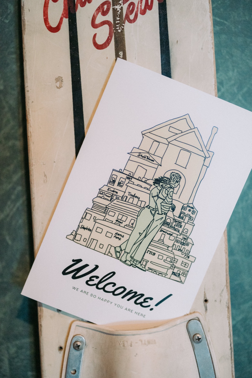 A hand-illustrated welcome card featuring the couple and a skyline rests atop a vintage water ski against a teal-painted wooden surface — Tim Larsen Photography, Brainerd Lakes MN
