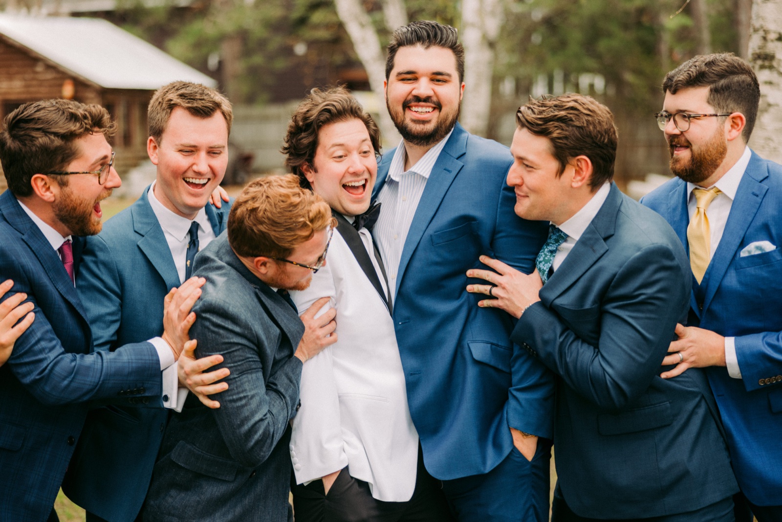 The groom laughs in the center of his groomsmen as they lean in around him in a tight, animated huddle outside the lodge — Tim Larsen Photography, Brainerd Lakes MN