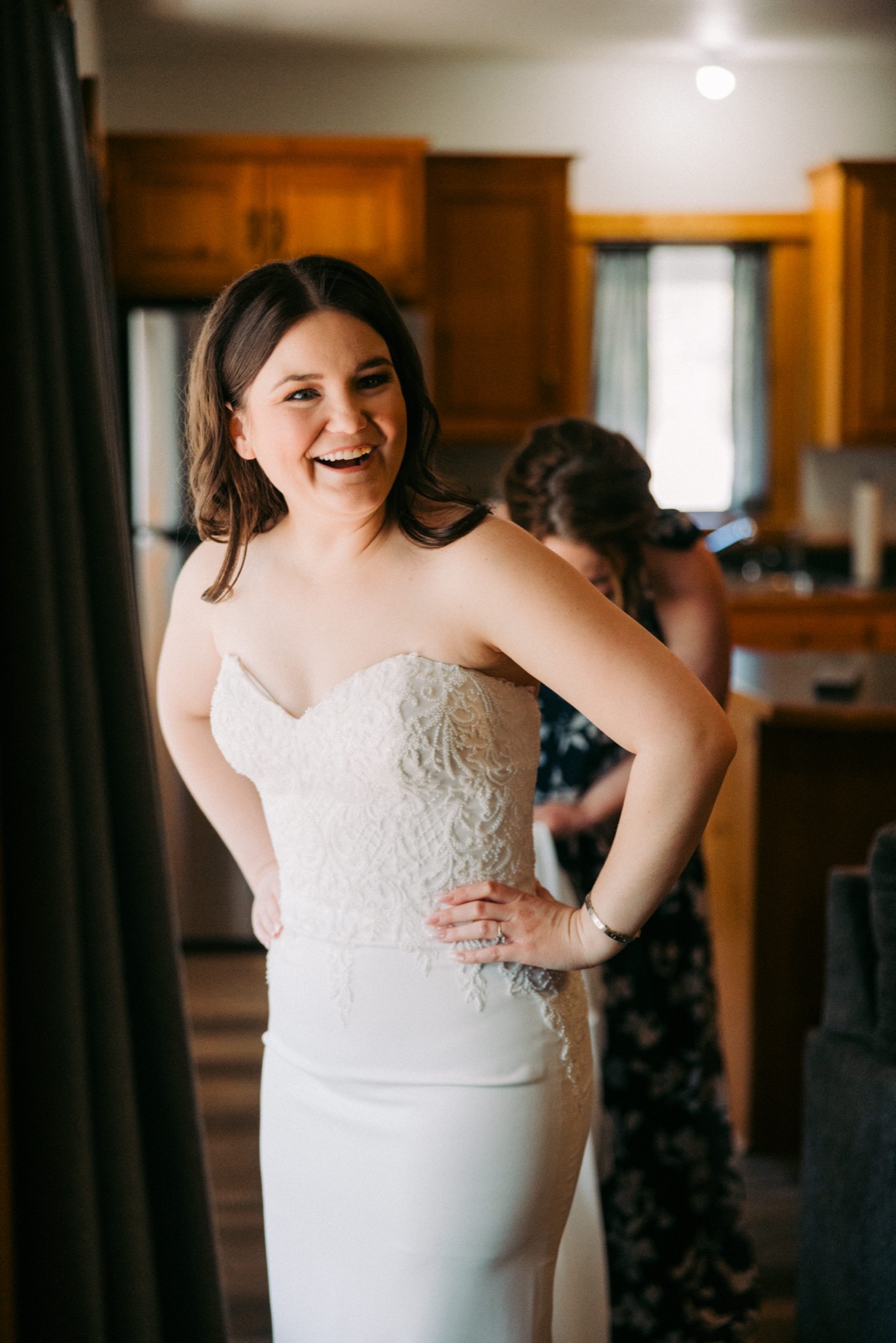 The bride laughs openly while a bridesmaid fastens the back of her lace strapless gown in a cabin kitchen — Tim Larsen Photography, Brainerd Lakes MN
