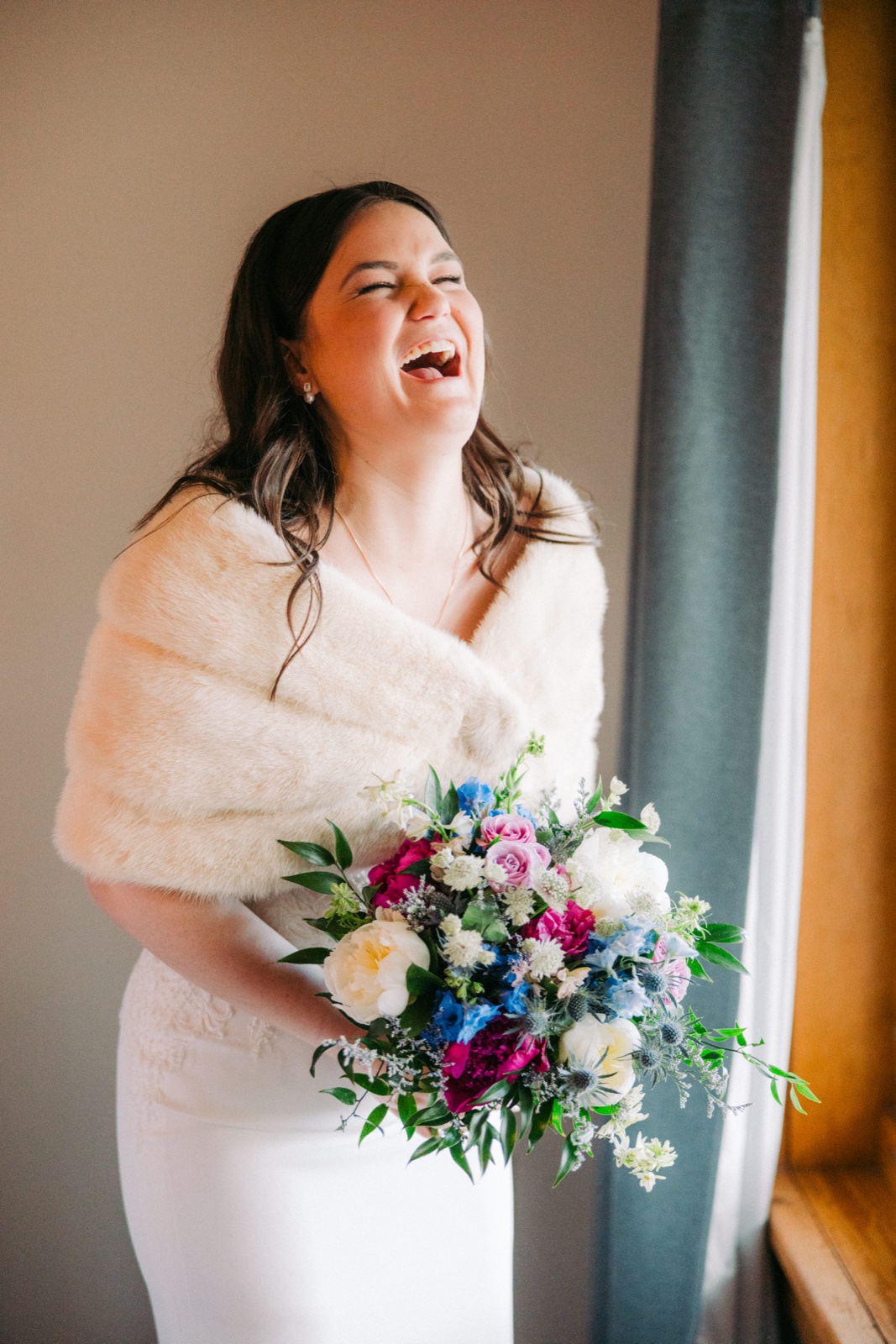 The bride laughs with her head thrown back while holding a vibrant blue, pink, and white bouquet, wrapped in a white fur stole beside a window — Tim Larsen Photography, Brainerd Lakes MN