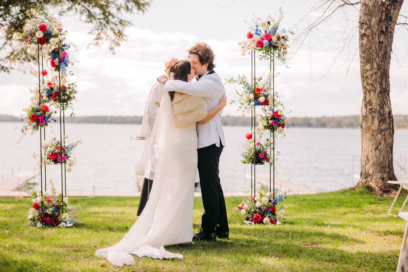 The bride and groom kiss between two tall pillar arrangements of bright florals while the officiant stands beside them, with a wide lake and bare-branched trees in the background — Tim Larsen Photography, Brainerd Lakes MN