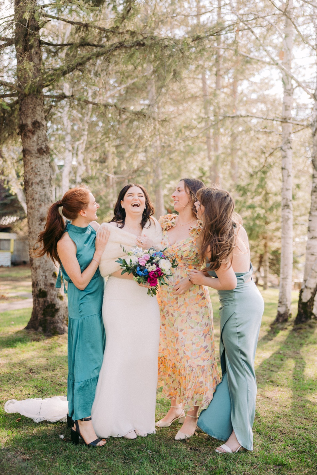 The bride laughs with her three bridesmaids in mismatched dresses among tall birch and pine trees, holding a colorful bouquet at Manhattan Beach Lodge — Tim Larsen Photography, Brainerd Lakes MN
