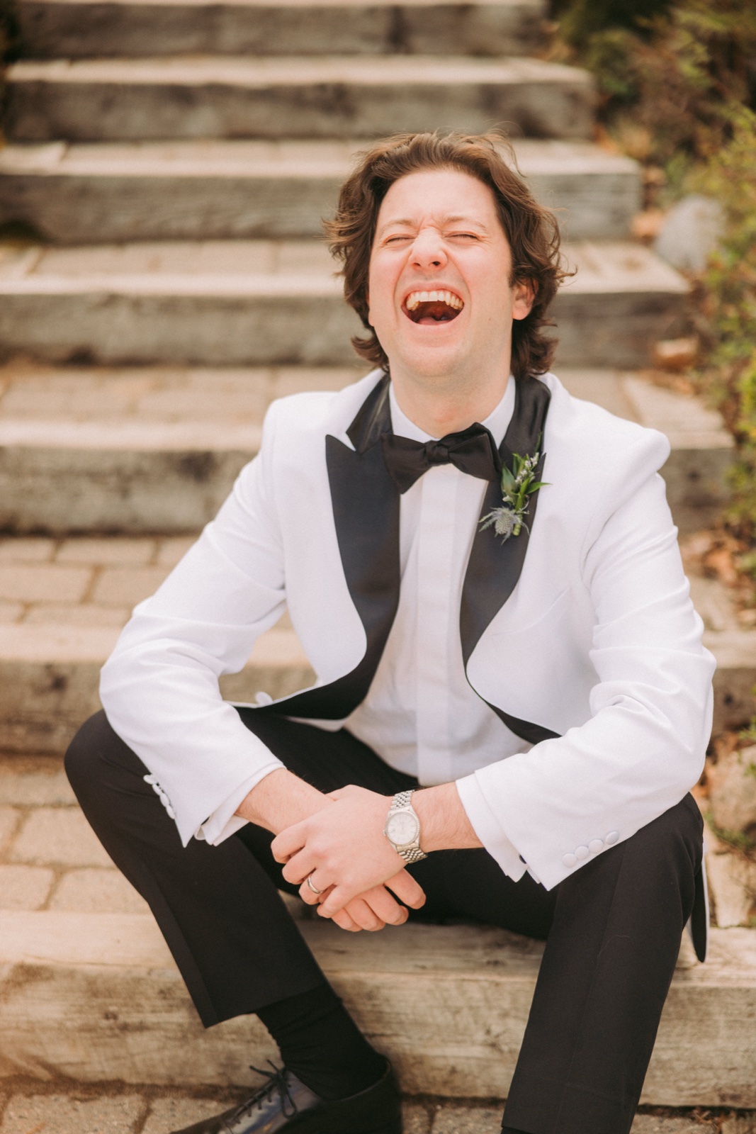 The groom in a white tuxedo jacket and black bow tie sits on stone steps, head tilted back in an open-mouthed laugh — Tim Larsen Photography, Brainerd Lakes MN