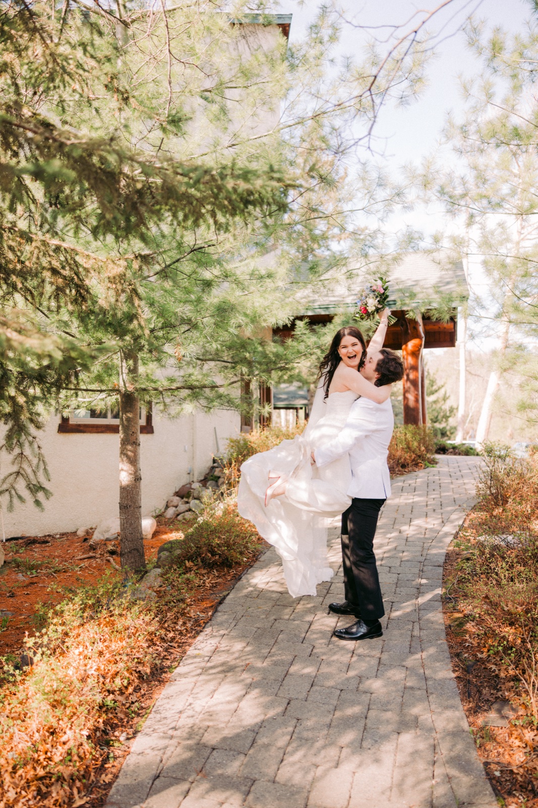 The groom lifts the bride off the ground as she raises her bouquet and laughs on a paver pathway outside Manhattan Beach Lodge — Tim Larsen Photography, Brainerd Lakes MN