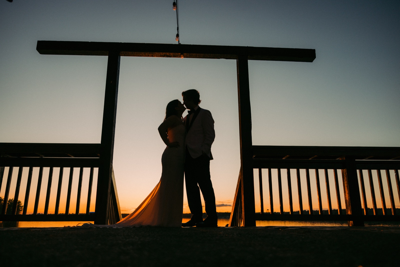 The bride and groom kiss in silhouette beneath the wooden lakeside arbor as the sky glows orange and blue at dusk over the water — Tim Larsen Photography, Brainerd Lakes MN