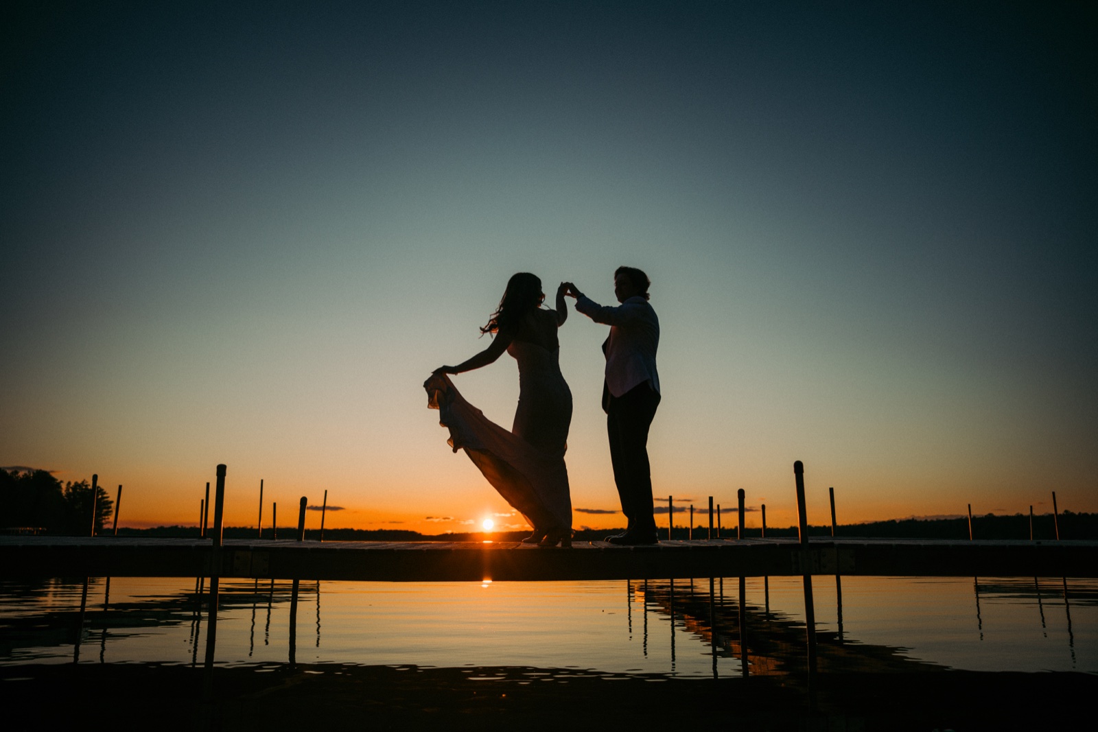 The bride and groom dance in silhouette on a long dock at sunset, the sun bursting between them and reflecting in the still lake below — Tim Larsen Photography, Brainerd Lakes MN