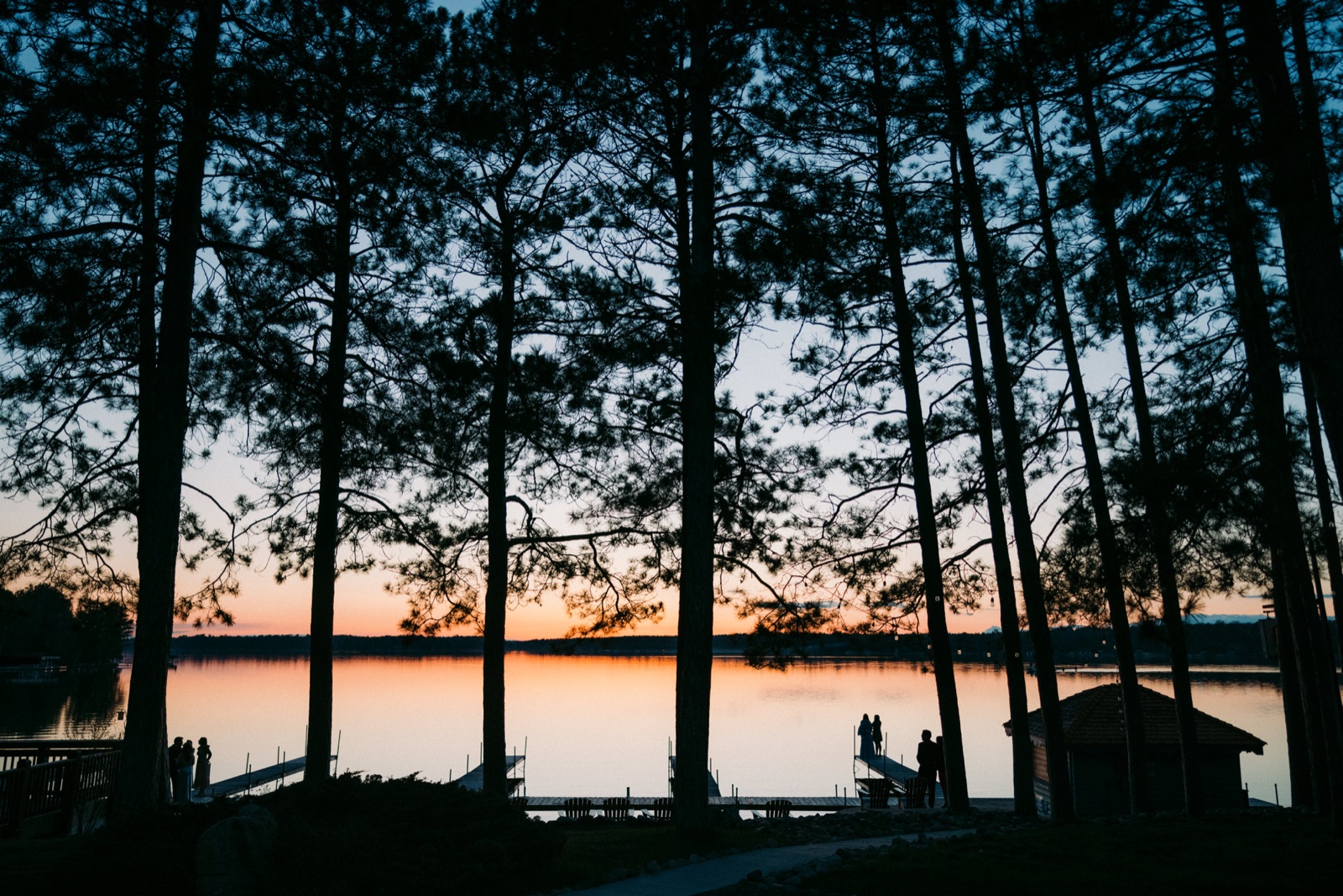 Towering pine trees silhouette against a peach and blue dusk sky over a calm lake, with small figures of guests and the couple gathered near the dock at Manhattan Beach Lodge — Tim Larsen Photography, Brainerd Lakes MN