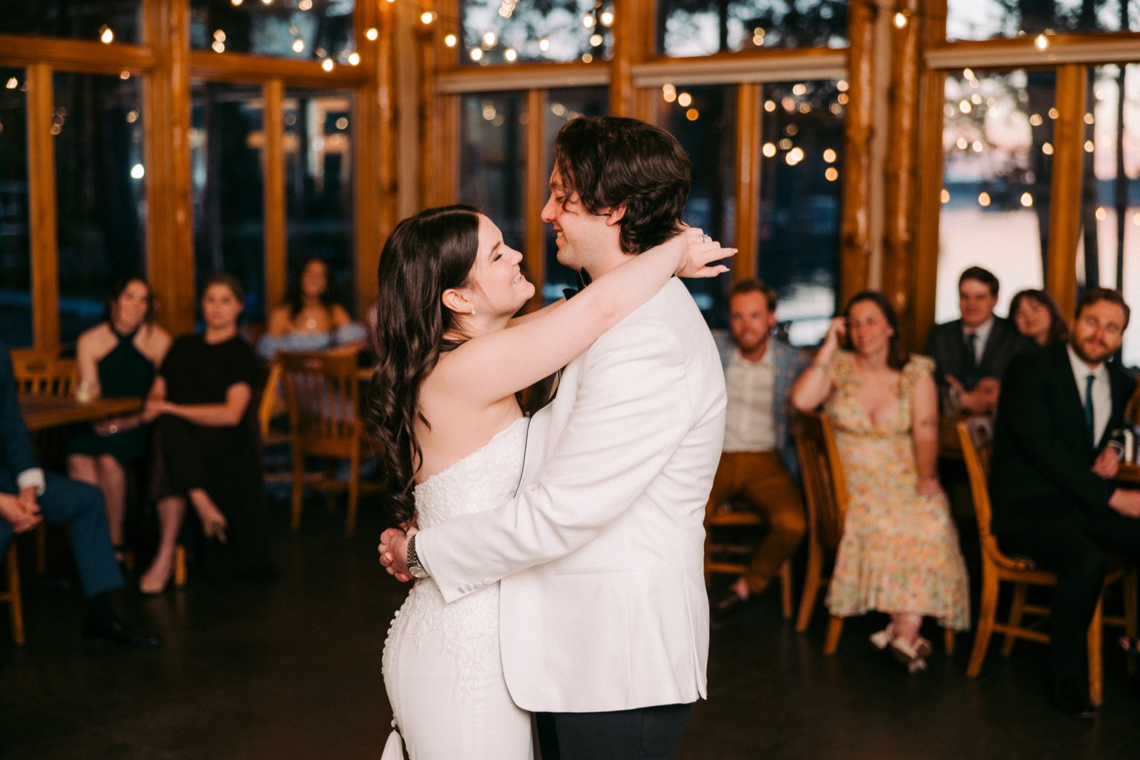 The bride smiles up at the groom as they hold each other on the dance floor, surrounded by seated guests inside the log-beamed lodge ballroom with the lake visible through the windows — Tim Larsen Photography, Brainerd Lakes MN
