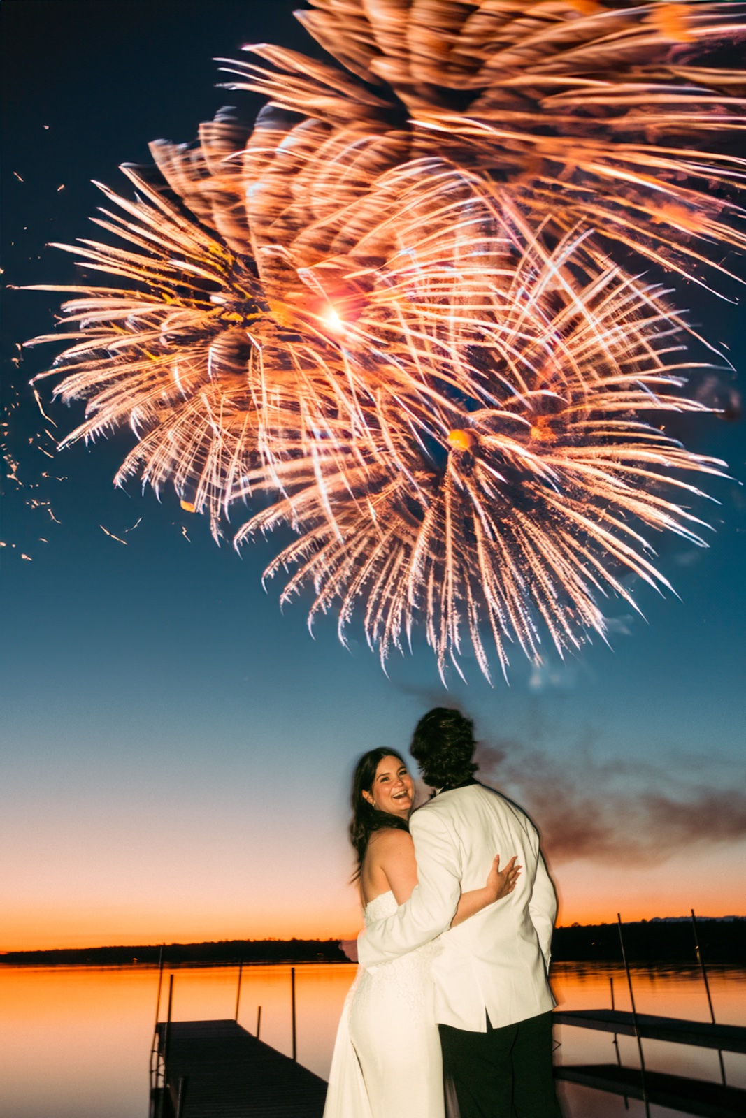 The bride beams over her shoulder while embracing the groom on a lakeside dock as a massive fireworks burst fills the dusk sky above them — Tim Larsen Photography, Brainerd Lakes MN