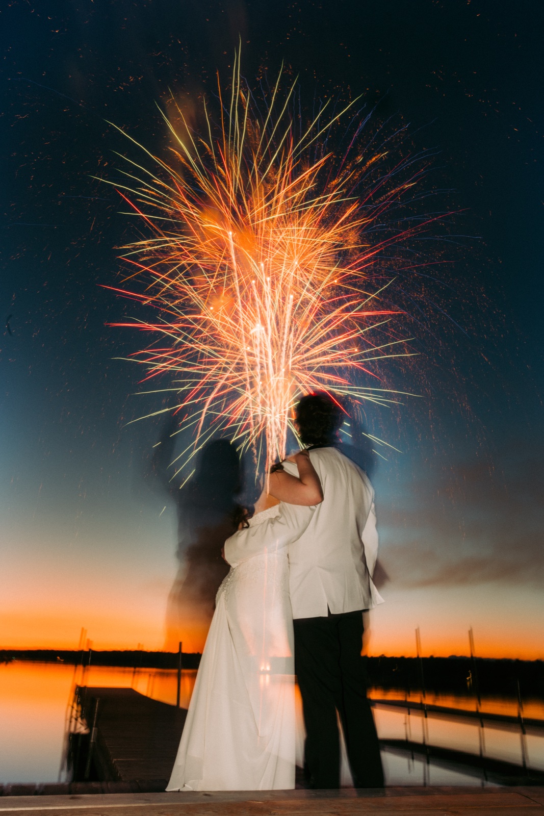 The bride and groom embrace on the lakeside dock at twilight while fireworks explode in the sky above them at Manhattan Beach Lodge — Tim Larsen Photography, Brainerd Lakes MN