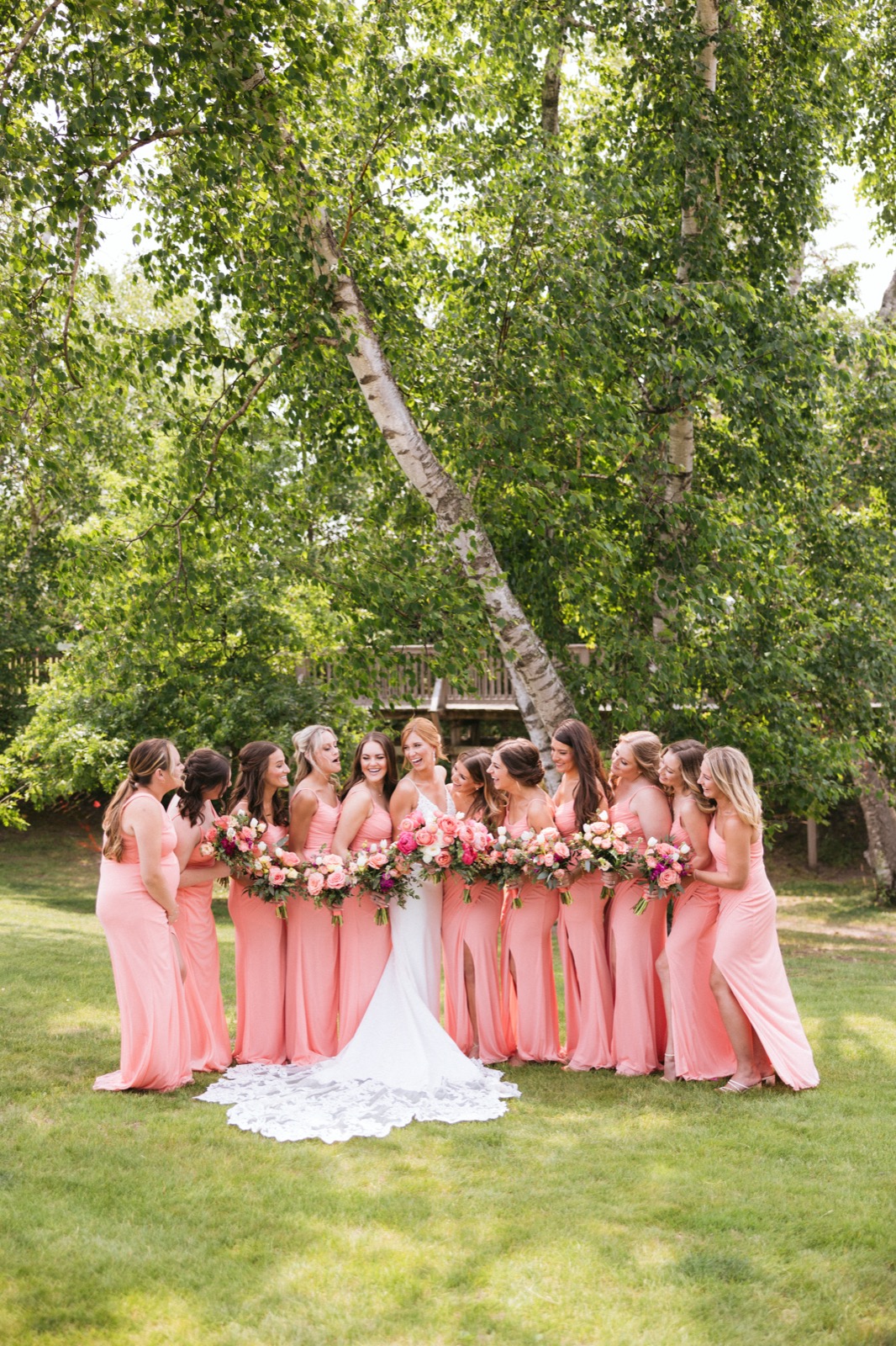 Melissa with ten bridesmaids in coral dresses laughing together on the lawn beneath a tall birch tree at Madden's — Tim Larsen Photography, Brainerd Lakes MN