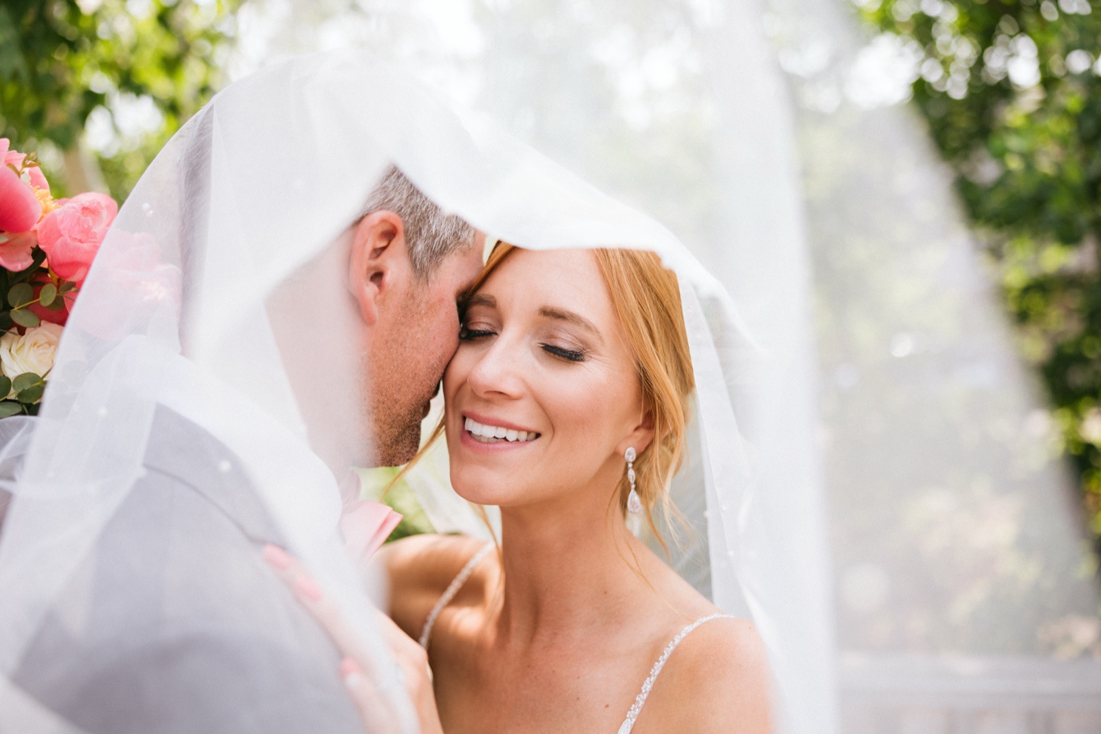 Melissa smiles with closed eyes as Kody presses his forehead to her temple, both framed beneath her translucent veil with coral peonies at the edge of the frame — Tim Larsen Photography, Brainerd Lakes MN