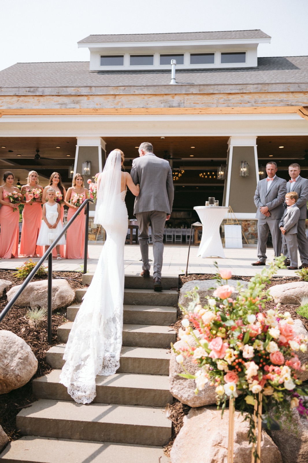 Melissa and her father walking up the stone steps toward the Madden's pavilion ceremony site, lined by coral-clad bridesmaids and gray-suited groomsmen — Tim Larsen Photography, Brainerd Lakes MN