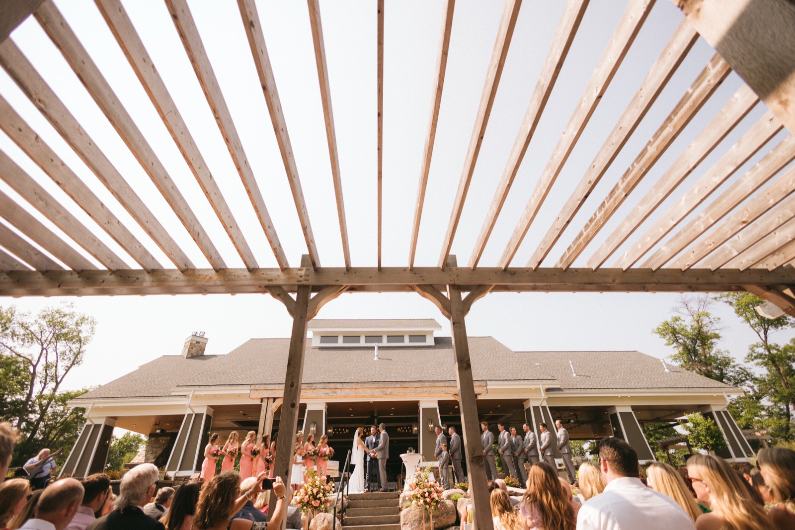 Wide low-angle view through radiating pergola beams of the outdoor ceremony at Madden's on Gull Lake, the bridal party gathered in front of the lodge — Tim Larsen Photography, Brainerd Lakes MN