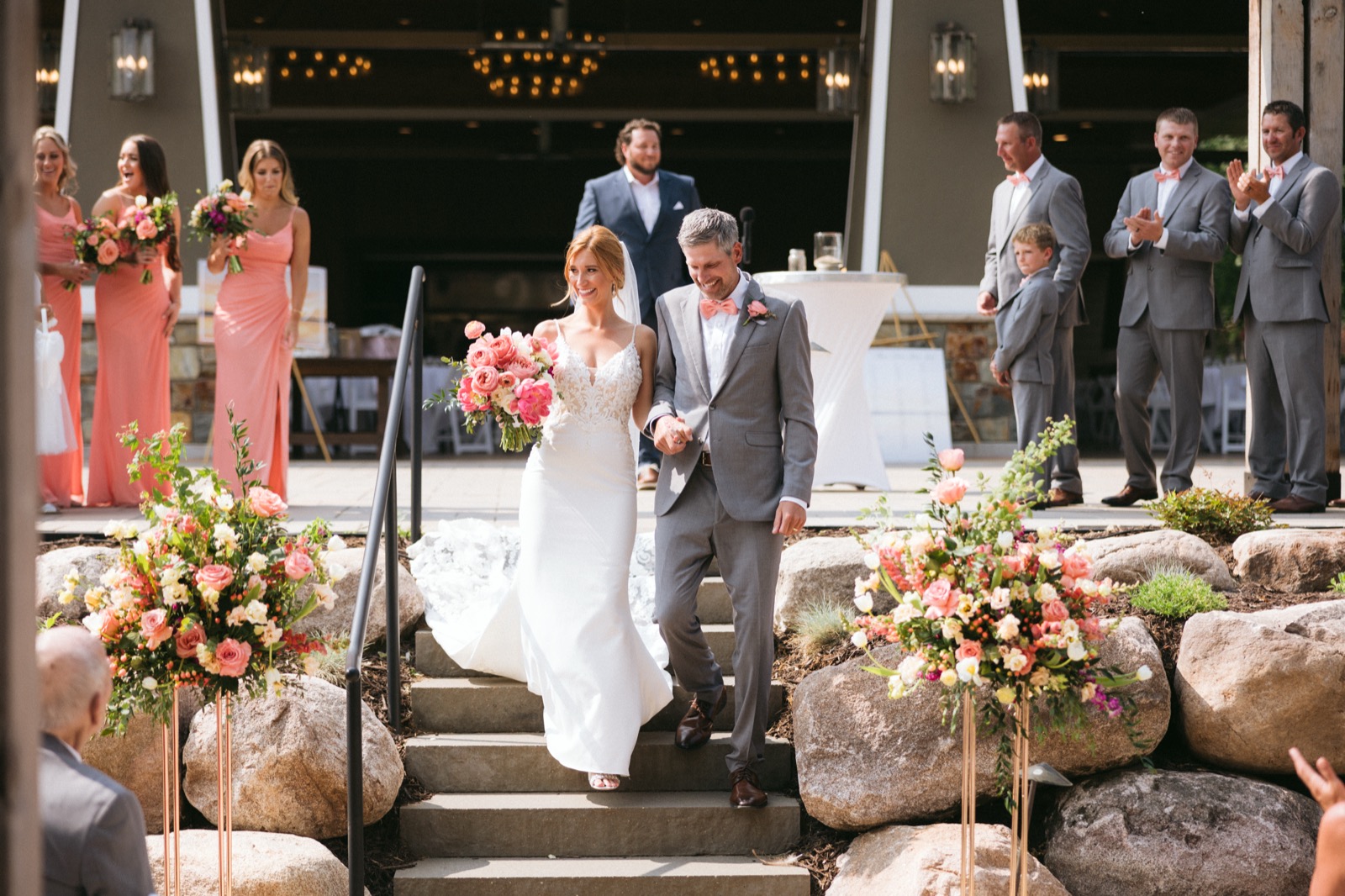 Melissa and Kody walking down stone steps lined with coral floral arrangements, their bridal party watching and applauding from the pavilion behind — Tim Larsen Photography, Brainerd Lakes MN