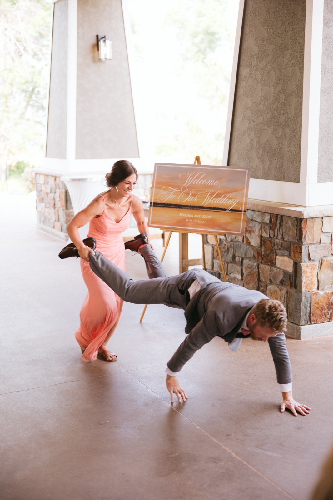A bridesmaid in coral holds Kody's legs as he walks on his hands past Melissa and Kody's wedding welcome sign on the covered Madden's walkway — Tim Larsen Photography, Brainerd Lakes MN