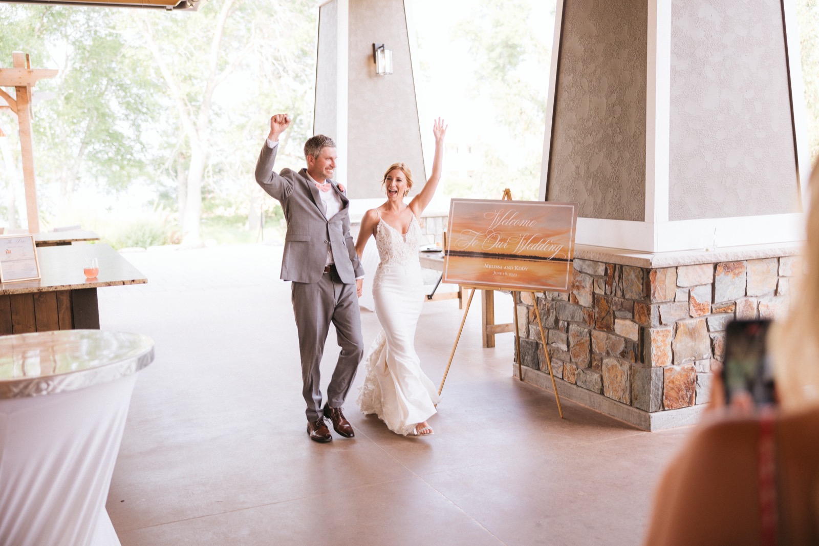Melissa and Kody framed between a stone pillar and their wedding welcome sign, arms raised mid-grand-entrance at the Madden's pavilion — Tim Larsen Photography, Brainerd Lakes MN