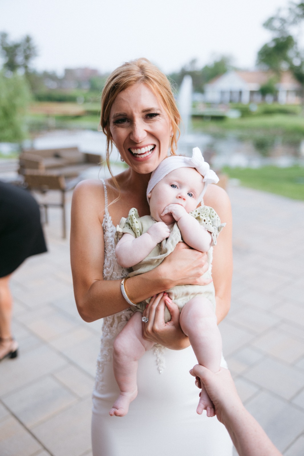 Melissa laughs widely as she holds a baby in a green romper and bow headband on the lakeside patio at Madden's, the lake and fountain just out of focus behind — Tim Larsen Photography, Brainerd Lakes MN
