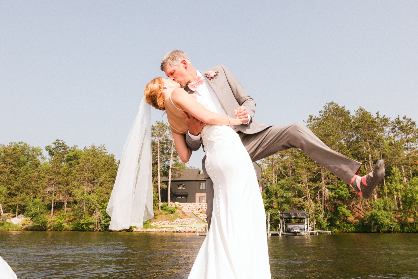 Low-angle dip-kiss on a Madden's dock against open sky, Melissa's veil trailing across the planks behind them — Tim Larsen Photography, Brainerd Lakes MN