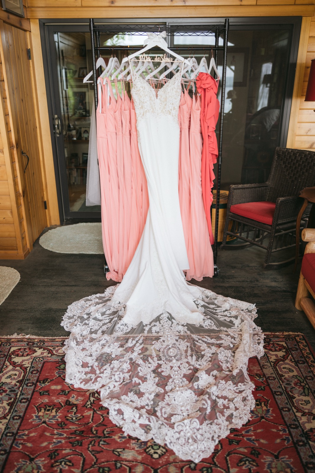 The bride's lace gown hanging centered on a rolling rack flanked by coral bridesmaid dresses, lace train fanned across a patterned rug in the Madden's cabin getting-ready room — Tim Larsen Photography, Brainerd Lakes MN