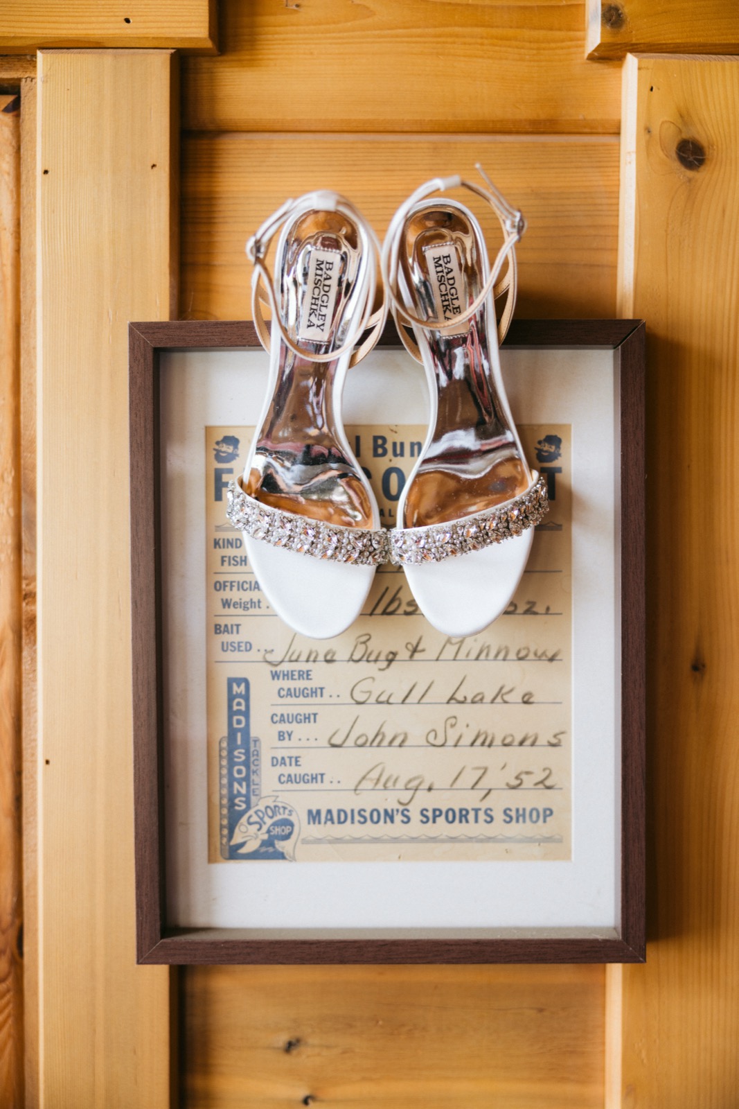 Crystal-strap bridal heels resting atop a framed 1952 Gull Lake fishing record on a knotty pine wall in the Madden's getting-ready cabin — Tim Larsen Photography, Brainerd Lakes MN