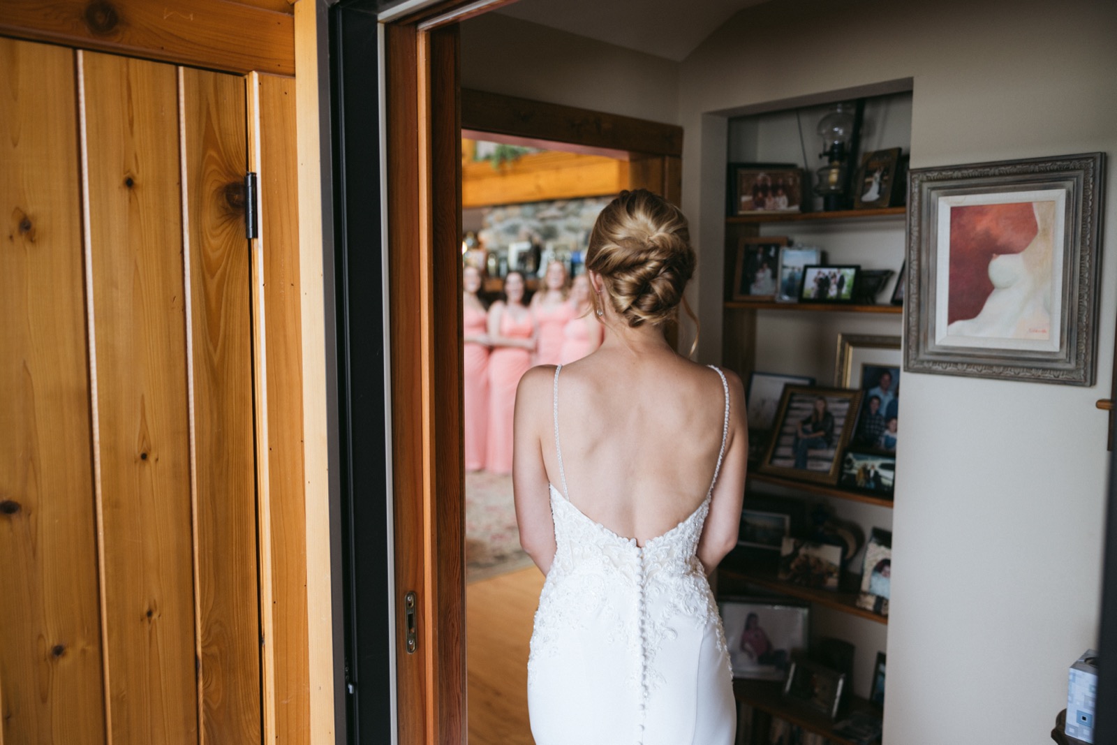 Bride seen from behind in a low-back lace gown walking through a wood-framed cabin doorway toward bridesmaids in coral waiting in the next room — Tim Larsen Photography, Brainerd Lakes MN