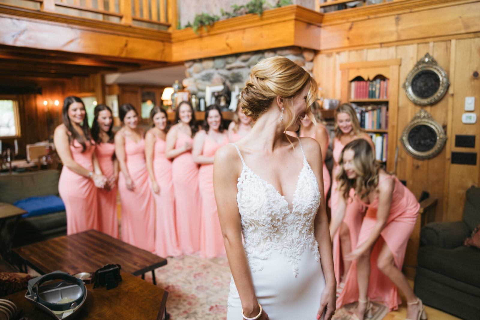 Melissa from behind as her bridesmaids in coral cluster around the stone fireplace in the lodge sitting room, reacting to the reveal at Madden's — Tim Larsen Photography, Brainerd Lakes MN