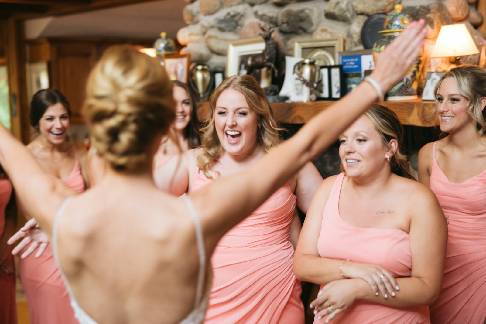 Bridesmaids in coral dresses react with open-mouthed delight as Melissa reveals her gown in the lodge living room with stone fireplace at Madden's — Tim Larsen Photography, Brainerd Lakes MN
