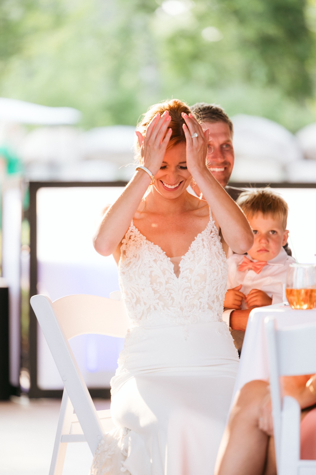 Melissa hands-to-face mid-laugh during toasts in the outdoor reception seating at Madden's, Kody and a young ring bearer watching from behind — Tim Larsen Photography, Brainerd Lakes MN
