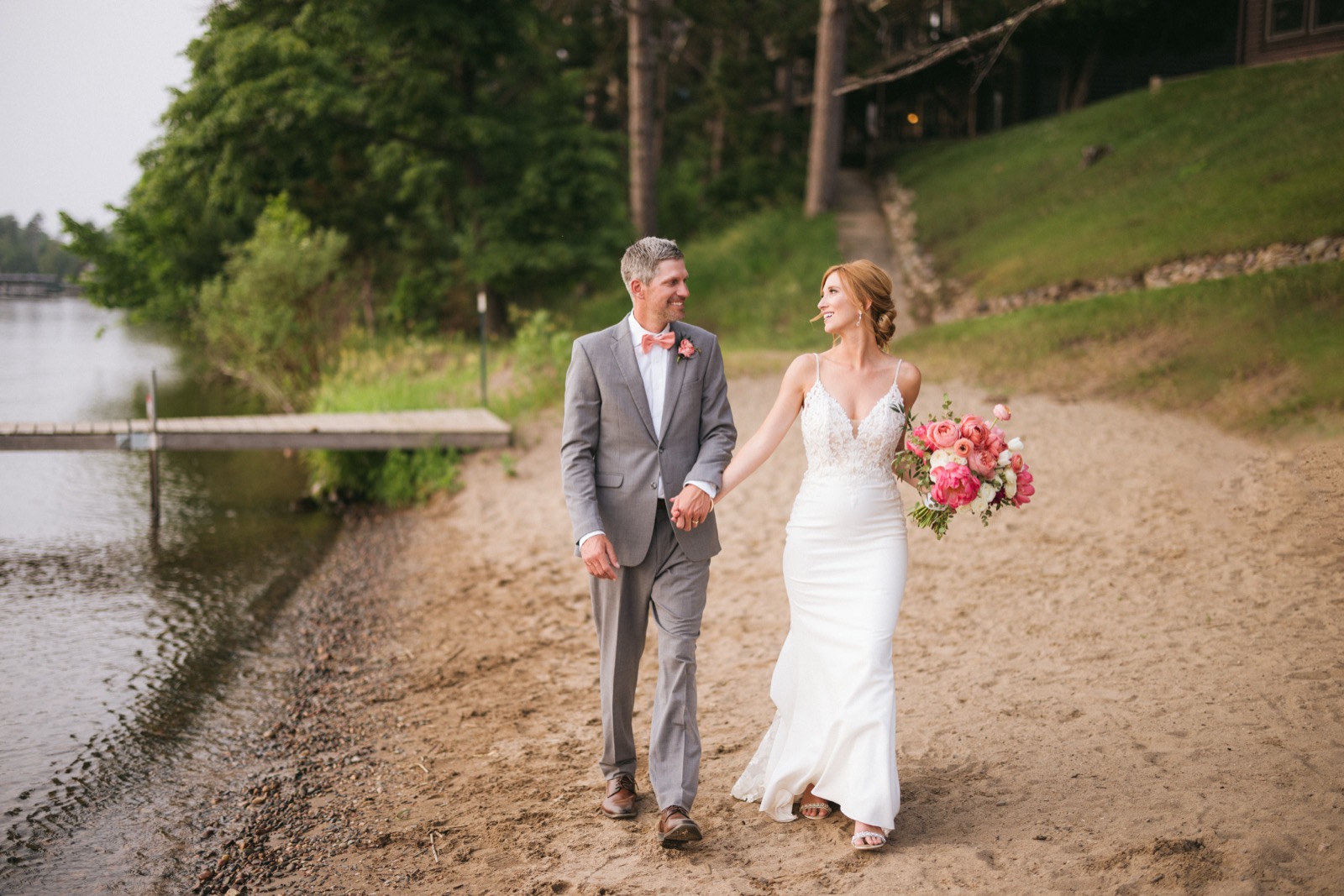 Melissa and Kody walking along the sandy lakeshore beach at Madden's, dock and treeline framing the lake at left — Tim Larsen Photography, Brainerd Lakes MN
